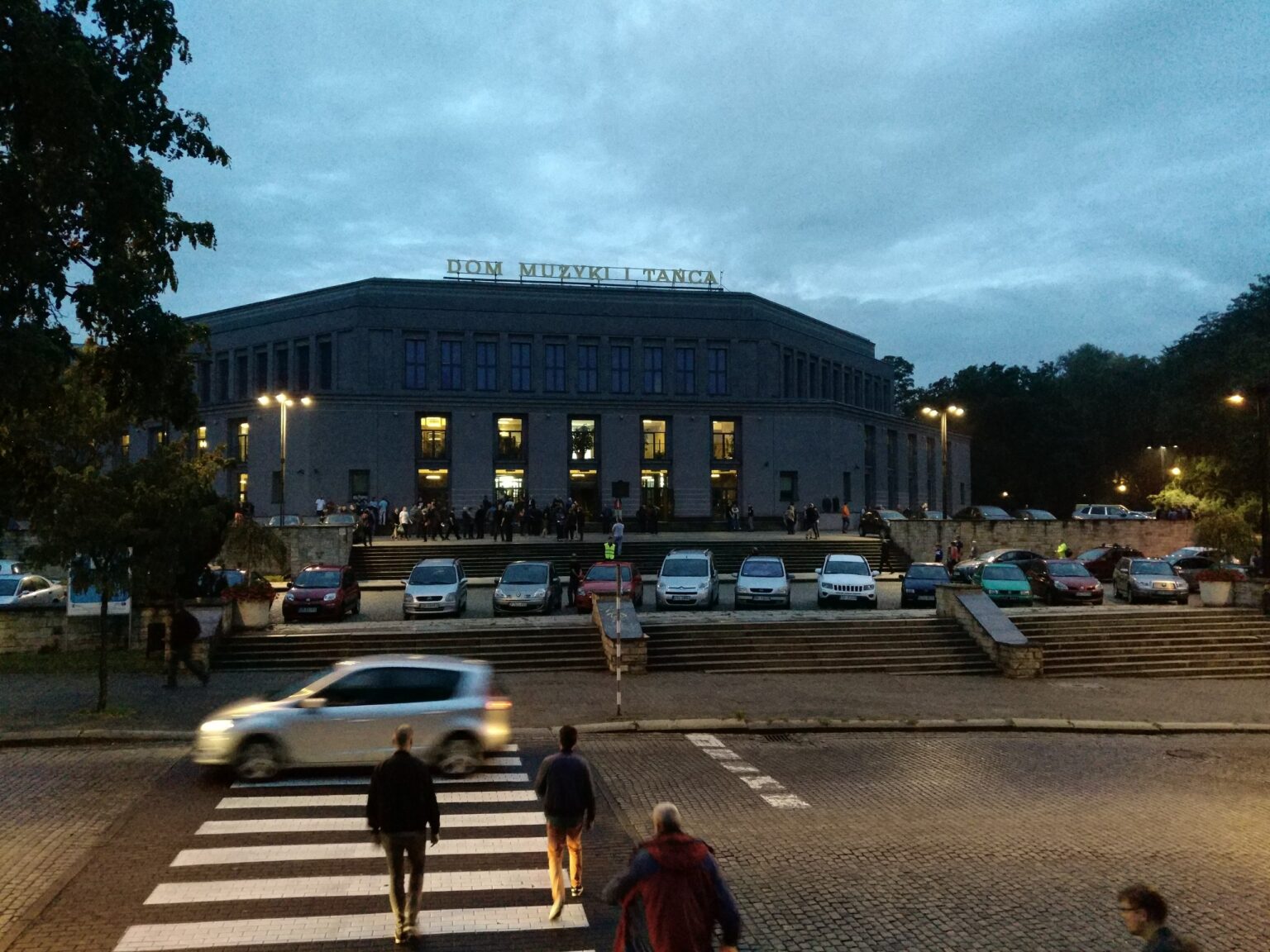 Modernistyczna bryła Domu Muzyki i Tańca w Zabrzu o zmierzchu, światła w oknach i rozmyty w ruchu samochód na pasach; reklamy Google Ads i Meta Ads dla firm z Zabrza